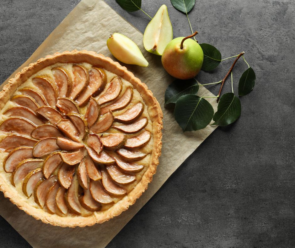 Une tarte aux poires posée sur une table avec 1 poire entière à côté et deux quartiers de poire